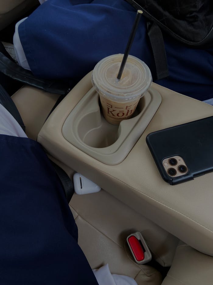Disposable coffee cup with straw in car cupholder beside smartphone on beige seat.