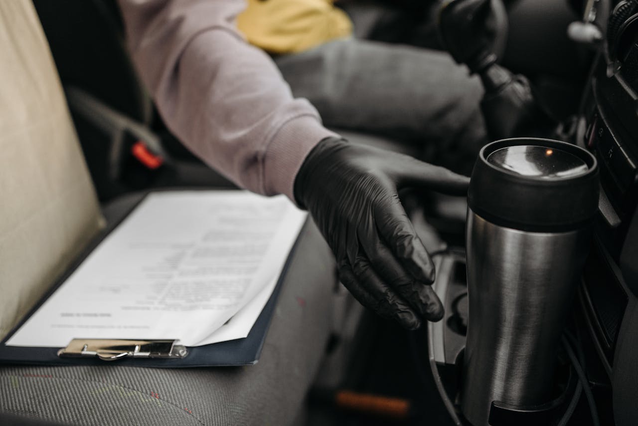 Close-up of a gloved hand reaching for a thermos in a car, with a clipboard on the seat.